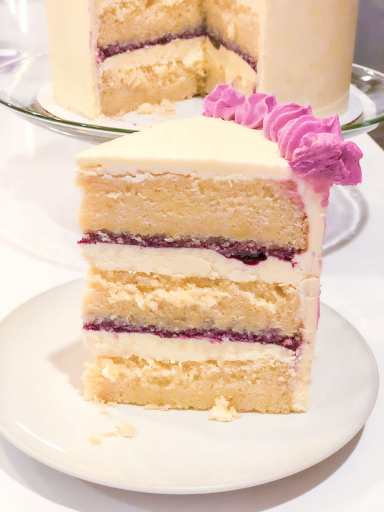 A cake slice in front of the Triple Layer Huckleberry and White Cake with White Chocolate Frosting.