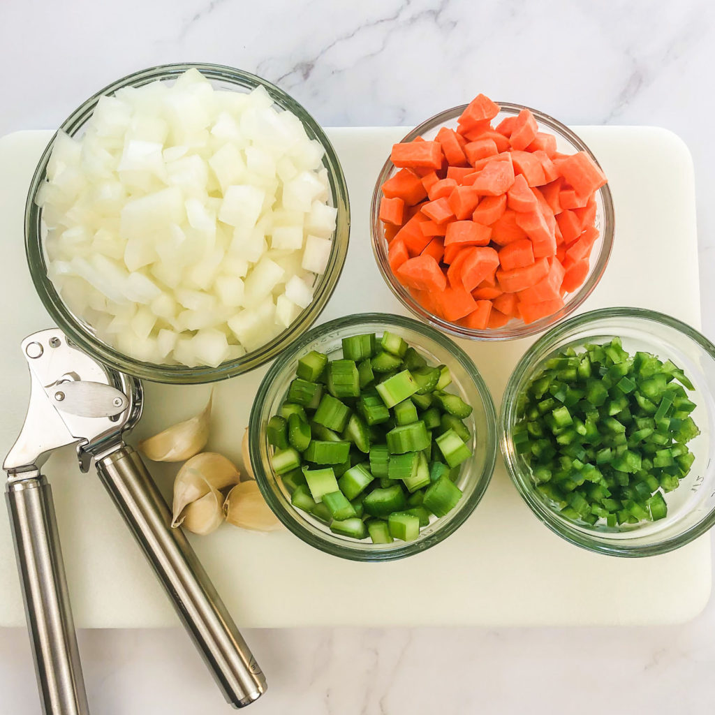 A cutting board with small bowls of chopped and prepped veggies.  A small bowl of chopped onions, a small bowl of chopped carrots, a small bowl of chopped celery, and a small bowl of diced jalapeño.  A garlic press and garlic cloves lay next to the bowls on the cutting board.