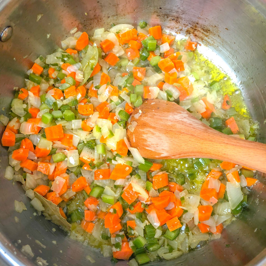 A pot with a wooden spoon stirring the sauteed carrots, onions, and celery.