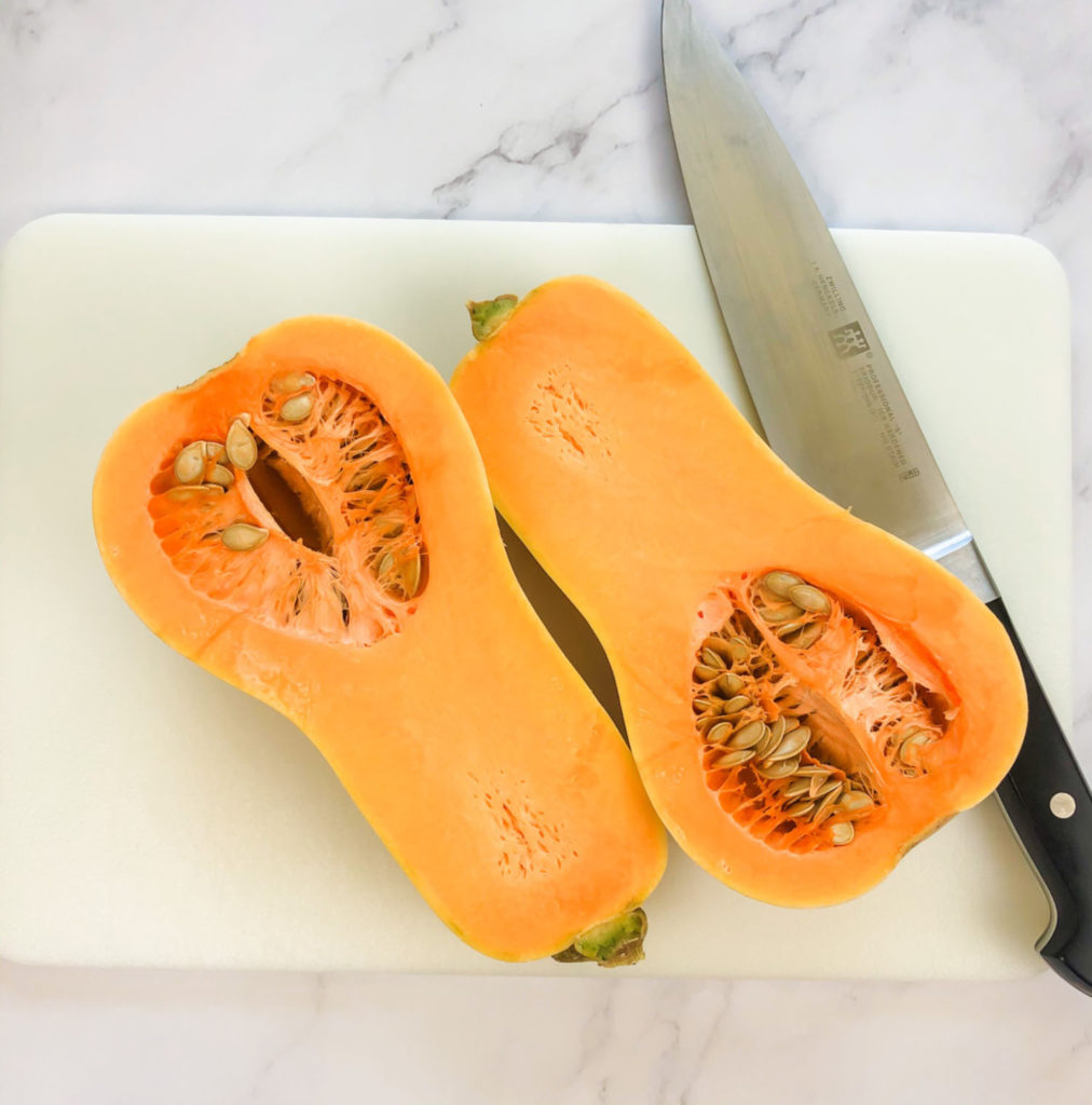 A butternut squash cut lengthwise on a cutting board.  The seeds have not been removed.  A knife lays next to the squash on the cutting board.