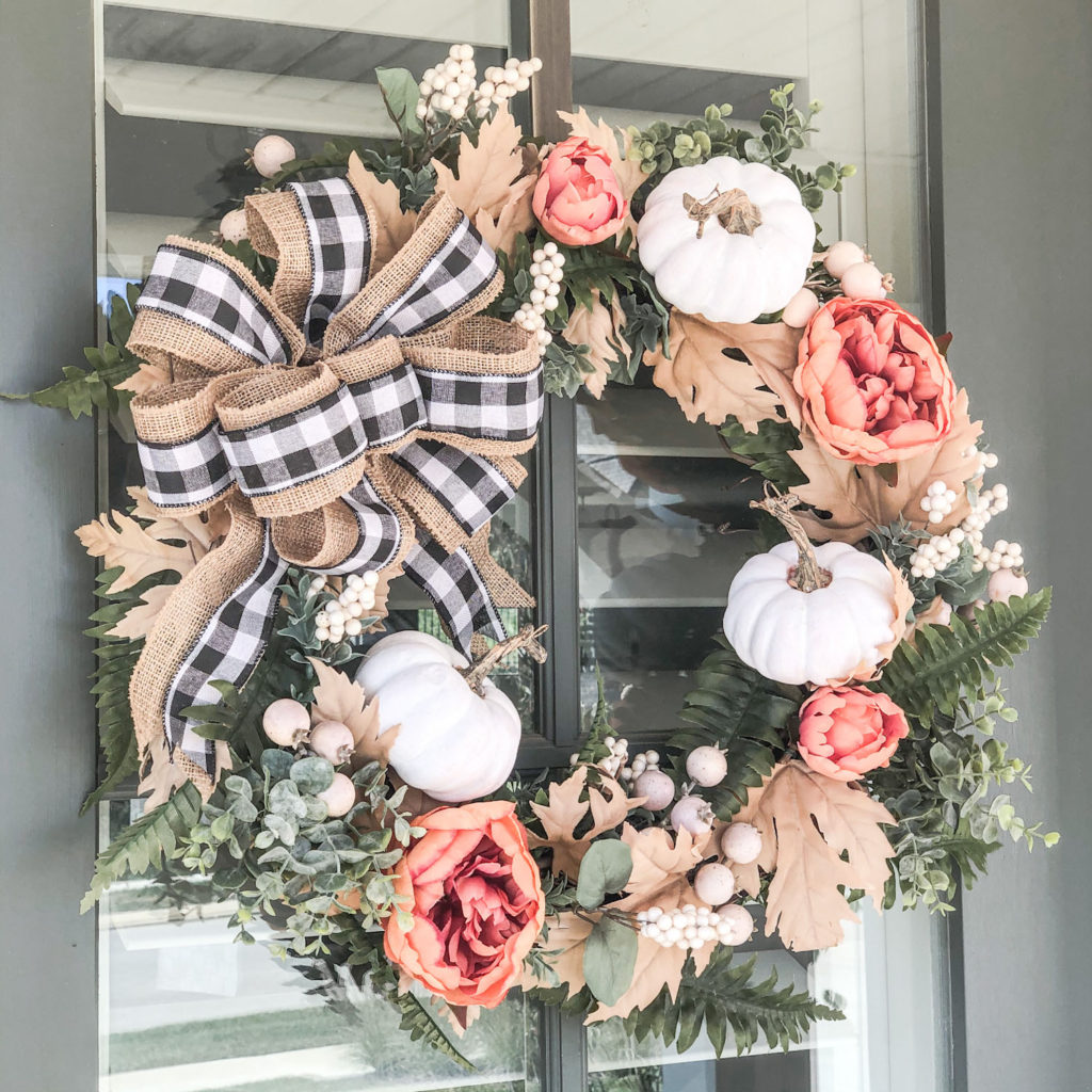 Beautiful DIY Farmhouse Fall Wreath displayed on a green door with windows.  Orange peonies, white berry picks, maple leaves, ferns, and eucalyptus are used to decorate the wreath.  A burlap and buffalo plaid ribbon is placed on the upper left corner.