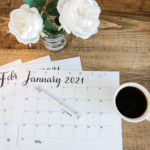 2021 free printable calendars on a desk. A cup of coffee sits to the right. White peony flowers in a vase sit on the desk.