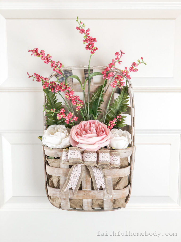 How to Make a Hanging Tobacco Basket for Valentine's Day displayed on a white interior door.  Pink and white peonies with pink, red, and white berry stem and fern leaves.  A burlap and pink lace bow is attached to the front of the tobacco basket.