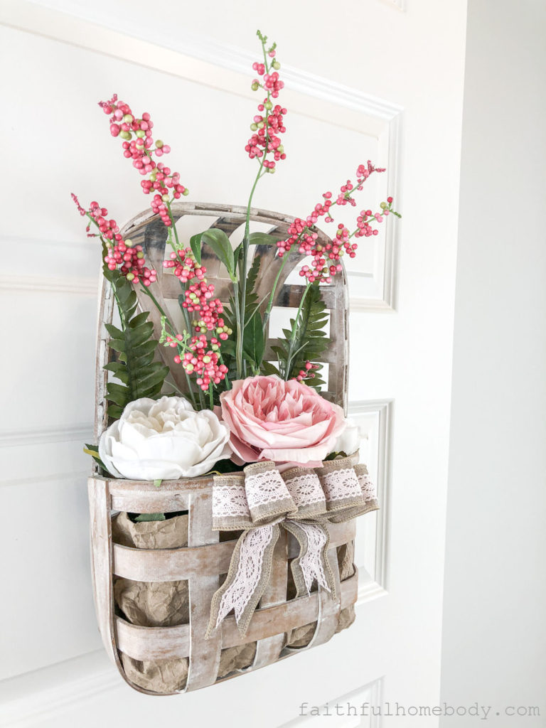 How to Make a Hanging Tobacco Basket for Valentine's Day.  Basket is displayed on a white interior door.  Pink and white peonies with pink, red, and white berry stem and fern leaves.  A burlap and pink lace bow is attached to the front of the tobacco basket.