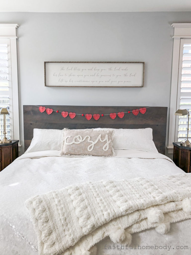2 Easy DIY Garlands for Valentine's Day.  Red Heart Doily Garland displayed on a headboard.  A pillow that says, "cozy", is on a white bedspread.