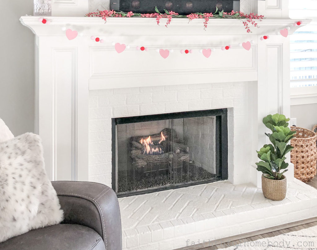 2 Easy DIY Garlands for Valentine's Day.  The pom-pom garland of light pink, red, and white poms and light pink paper hearts are displayed on a mantle.  A fire is in the fireplace.  A green fiddle leaf plant sits on the hearth.  