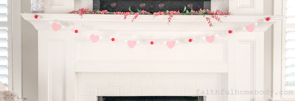 Pom pom garland with pink paper hearts. Pink, red, and white pom poms. The garland is stretched across a mantle. Pink floral berries are layed on the mantle above where the garland is displayed.