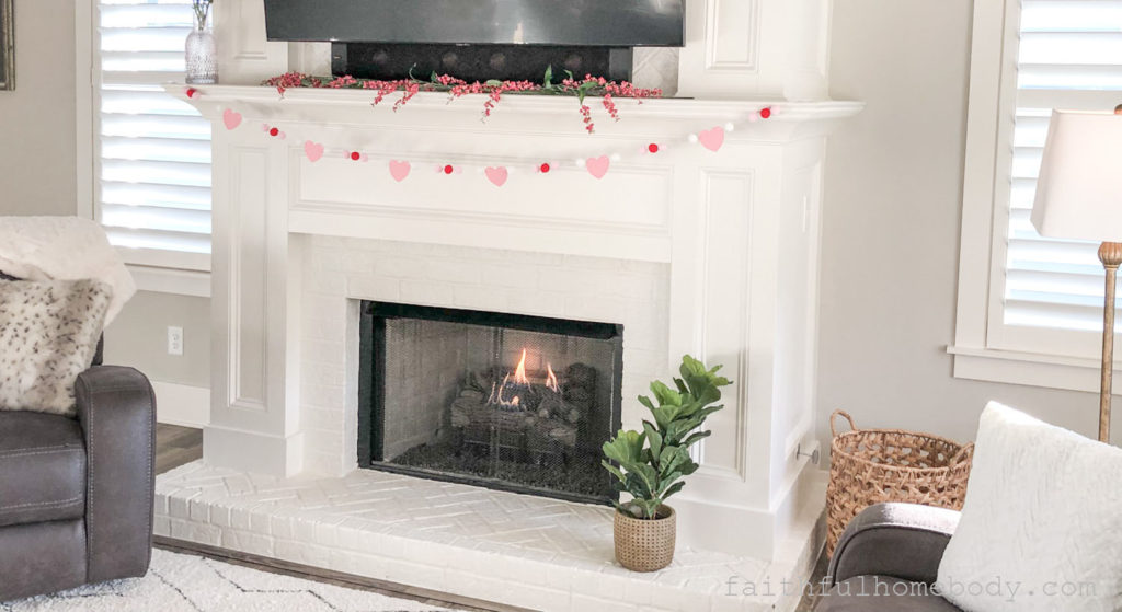2 Easy DIY Garlands for Valentine's Day.  The garland is displayed on a white mantel with a fire going in the fireplace.  A plant sits on the hearth.  A basket is beside the fireplace.  Two windows with shutters are on either side of the fireplace.
