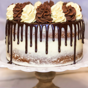 A double 8 inch Boston Cream Pie on a cake stand. The cake stand has a scalloped white rim and light wooden turned base. The cake is crumb-coated with vanilla buttercream frosting. Dark chocolate ganache drip on the top and sides. Topped with chocolate buttercream swirls and vanilla custard swirls.
