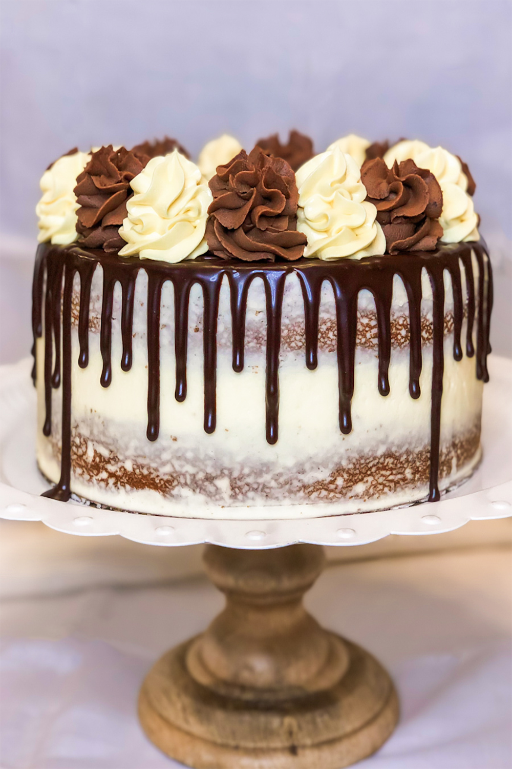 A double 8 inch Boston Cream Pie on a cake stand.  The cake stand has a scalloped white rim and light wooden turned base.  The cake is crumb-coated with vanilla buttercream frosting.  Dark chocolate ganache drip on the top and sides.  Topped with chocolate buttercream swirls and vanilla custard swirls. 