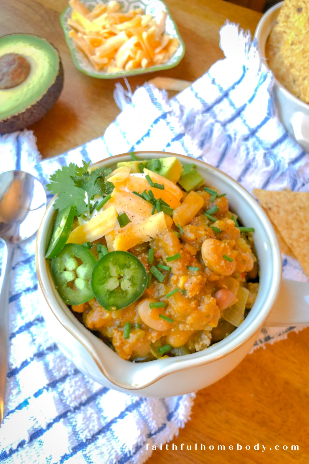 A white bowl contains Slow-Cooker Turkey and Two-Bean Pumpkin Chili..  The chili is topped with sour cream, shredded cheddar cheese, chives, sliced jalapenos, and cilantro.  The bowl is sitting on a white and blue kitchen towel with tortilla chips , avocado, and a dish with shredded cheese are in the background.