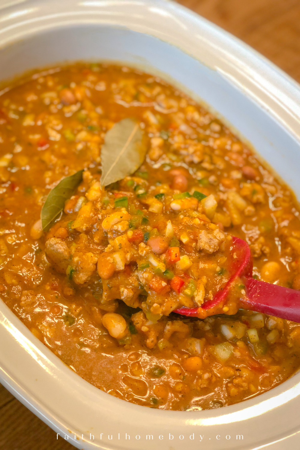 A white slow-cooker is filled with the Turkey and Two-Bean Pumpkin Chili.  A red spatula is dipped into the chili, emphasizing the ingredients.